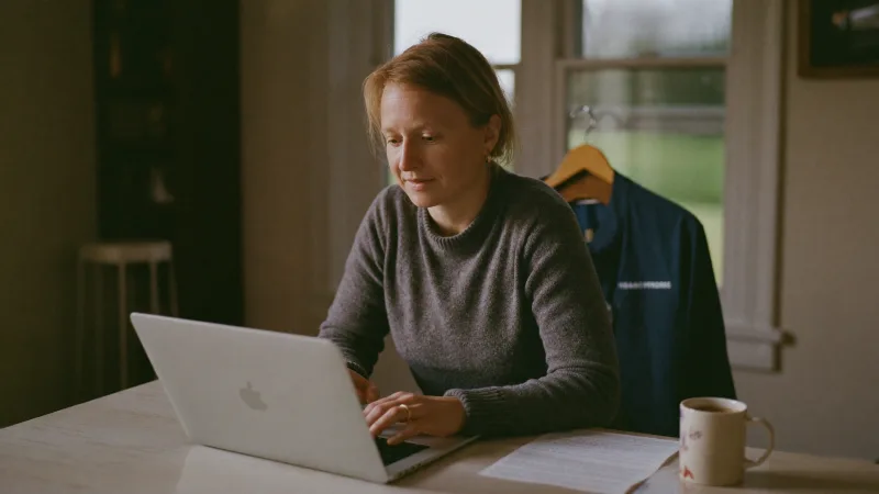 A healthcare worker updating her CV on a laptop at a kitchen table, uniform hanging on the chair behind her