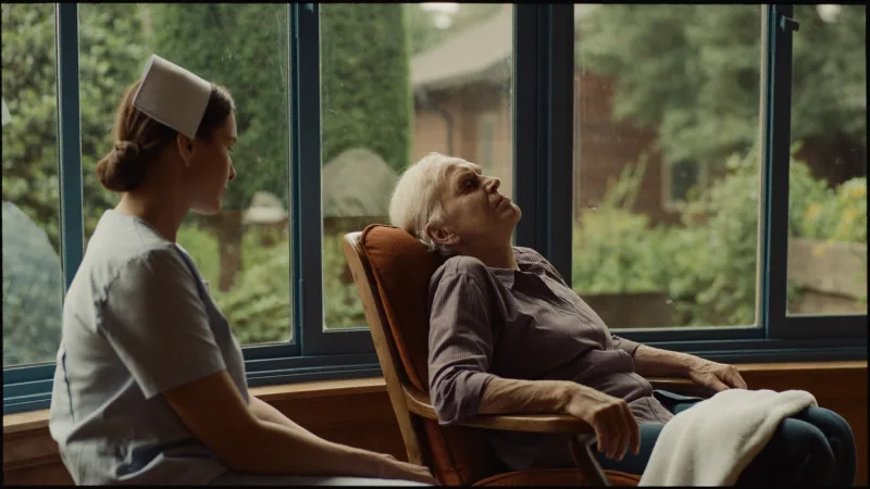 A hospice nurse sitting with a patient beside a window looking out onto a garden