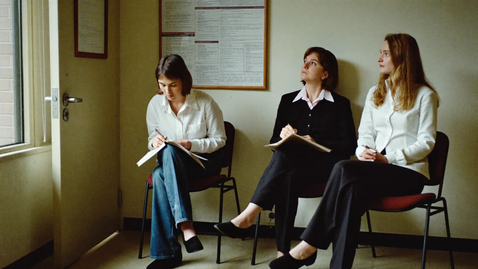 Candidates waiting on chairs outside an interview room in a hospital corridor