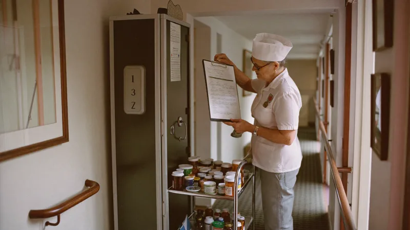 A senior carer preparing a medication round from a locked trolley in a care home corridor